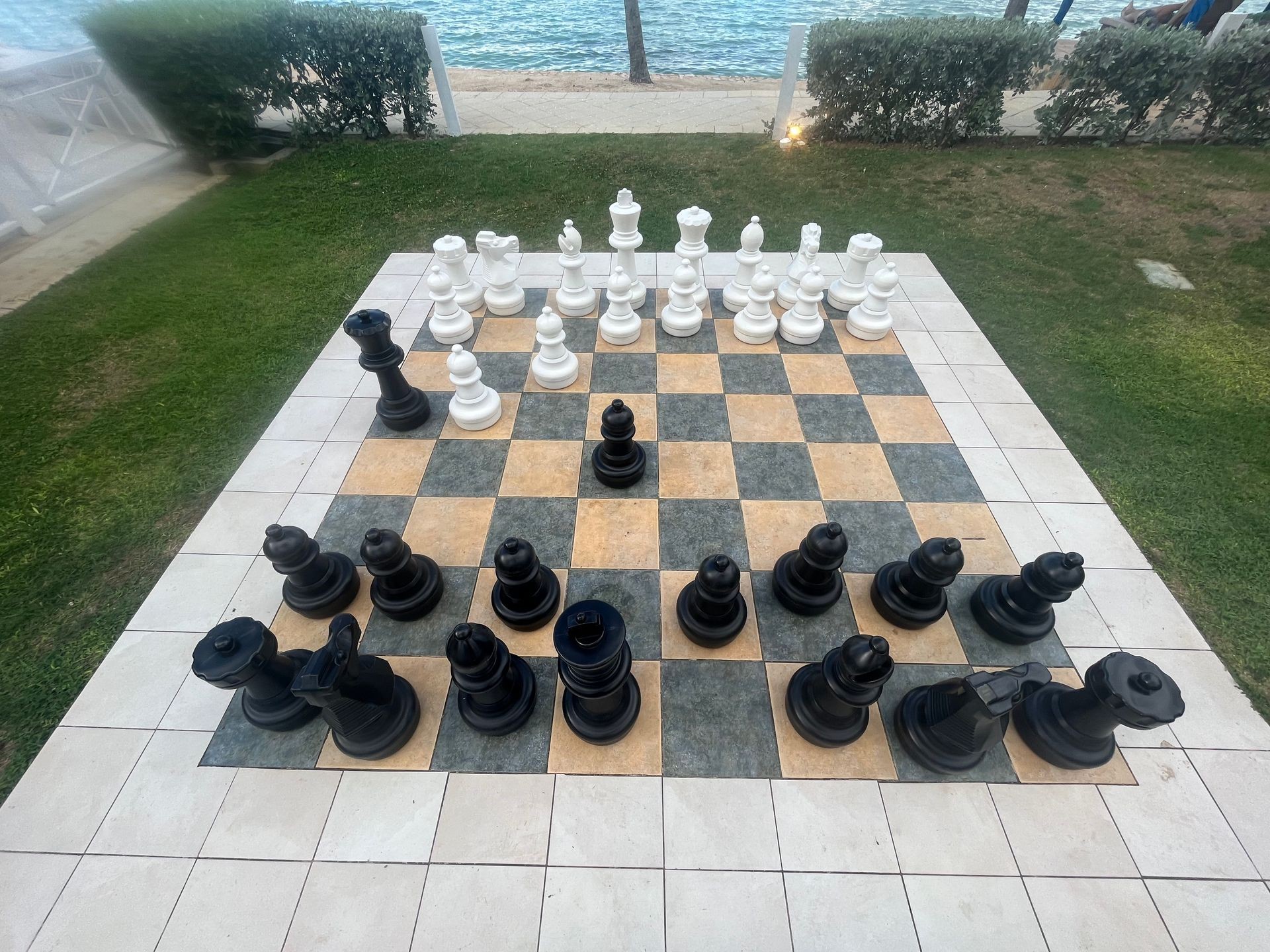 Large outdoor chessboard with oversized black and white chess pieces positioned on a grassy area near water.